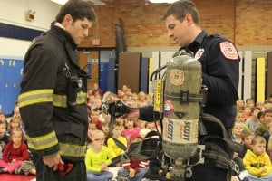 Firefighters Ian Ballard (in turnout gear) and Eric Stanley work together to put the air pack on Ballard.