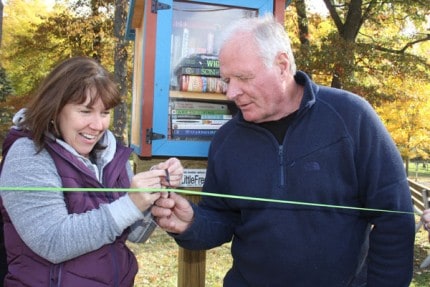 Sue Whitmarsh, president of the Friends of the Library and Gary Muncie, who helped plant the little library, cut a little ribbon with little scissors in front of the Little Free Library in Pierce Park.